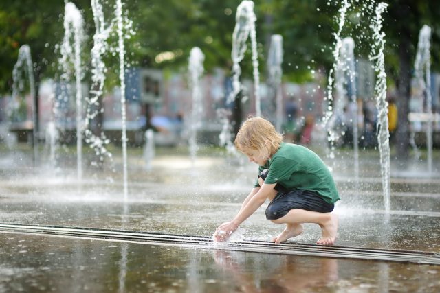 Little boy plays in the square between the water jets in the city fountain at sunny summer day. Active summer leisure for kids in city.